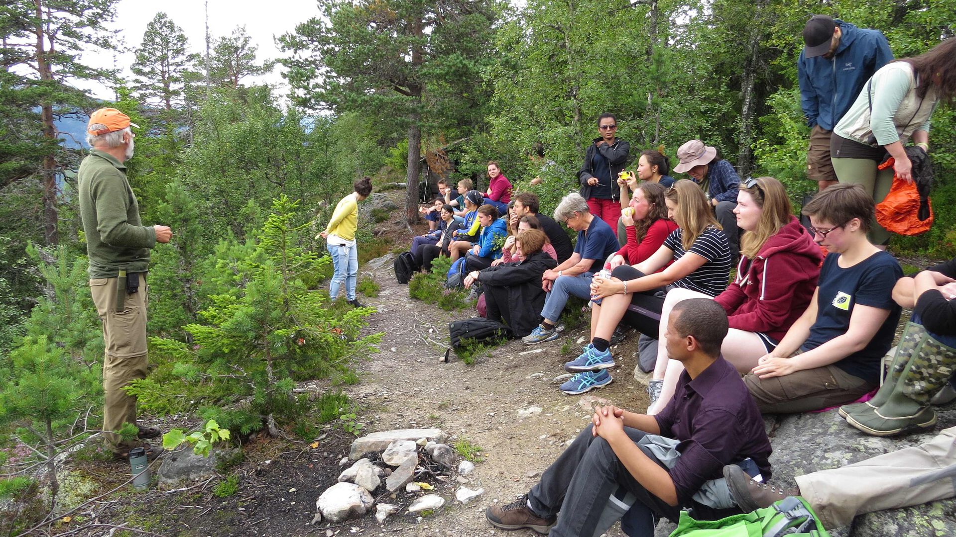 A group of students are sitting in the forest. In the background there is green trees. The teacher is standing infront of the students talking.