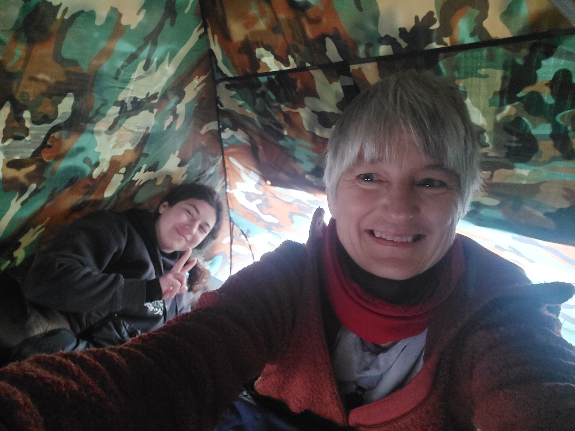 A teacher and a student in a tent with camouflage-pattern. The teacher smiles, and the student in the background smiles and shows the "peace" sign.