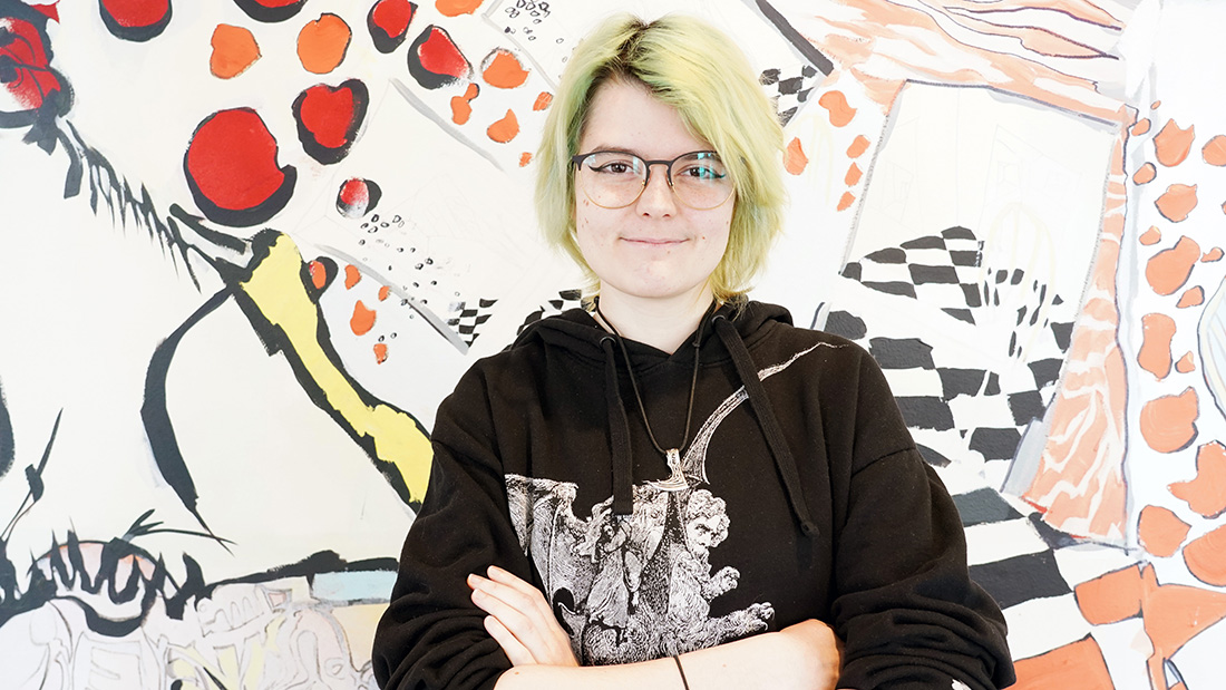 Photo of a female student in front of an artwall. She has yellow hair, glasses and a black hoodie. 