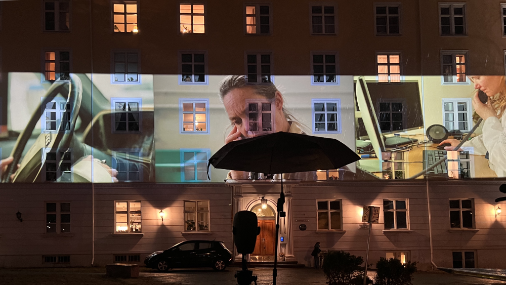 A triptych of videos is projected onto the façade of a house at night. The leftmost shows hands on the steering wheel of a car, the middle a woman in a phone conversation, and the rightmost another woman at a hospital office having a phone conversation. In front of the building, we see the black silhouette of a microphone shaped as a dummy head, protected by an umbrella.