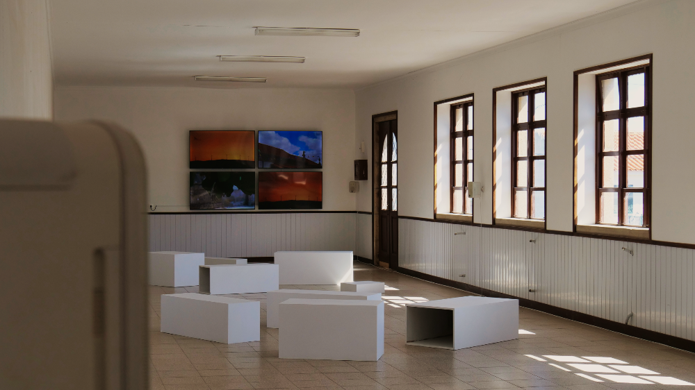 A room in a former school building in the village of Poço do Canto. Four videos on the wall show videos filmed in the surroundings, and plinths in the room offer seating for the audience. Part of a loudspeaker can be seen in the front.