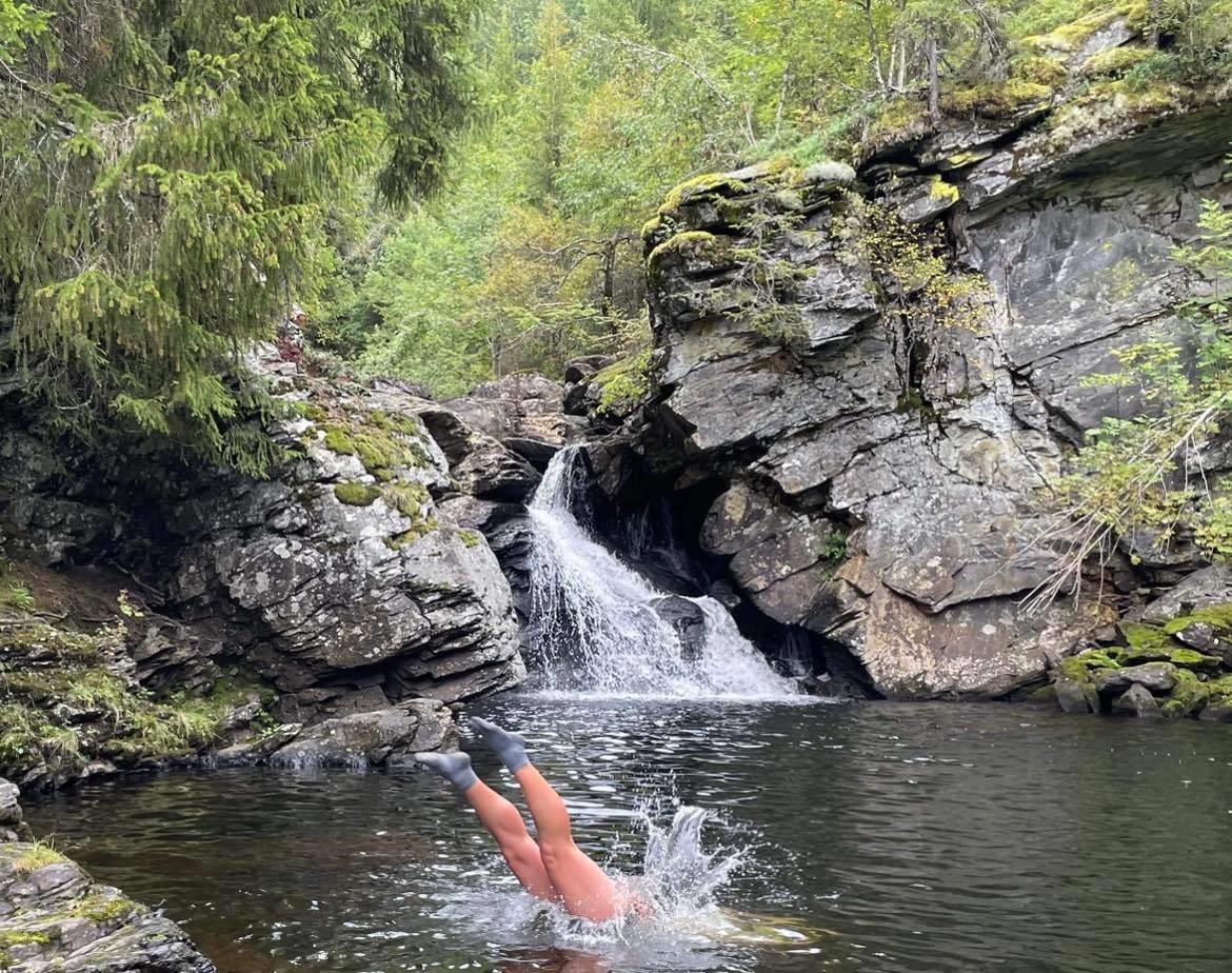 person diving in a lake in front of a waterfall, only legs and feet sticking up from the water
