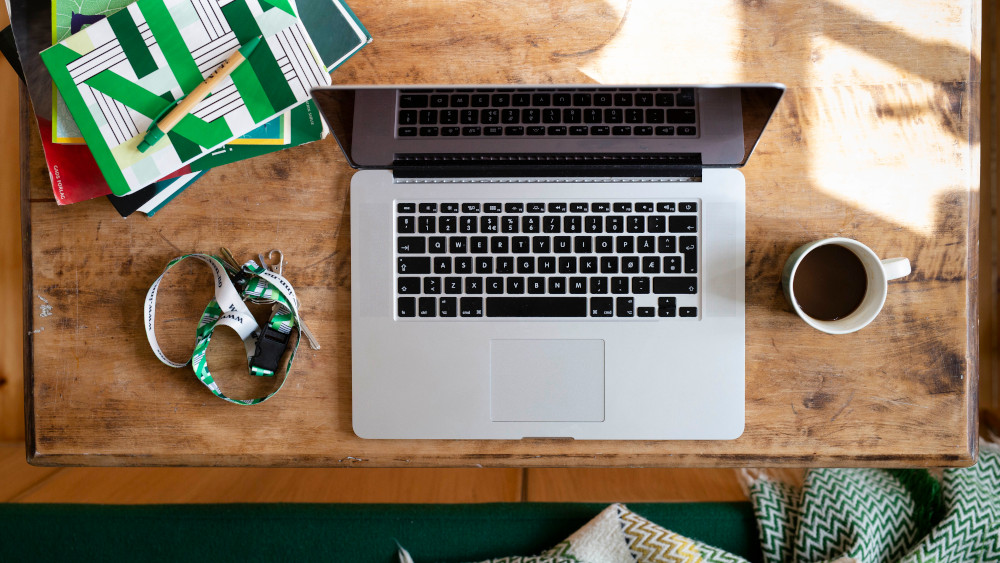 En laptop, kaffekopp, notatbok, penn og nøkkelknippe i bånd ligger på et skrivebord