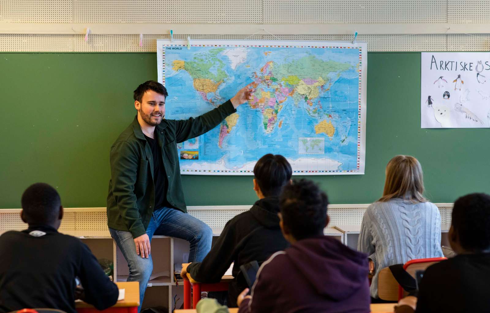 Mannlig lærer peker på kart foran elever i klasserom.