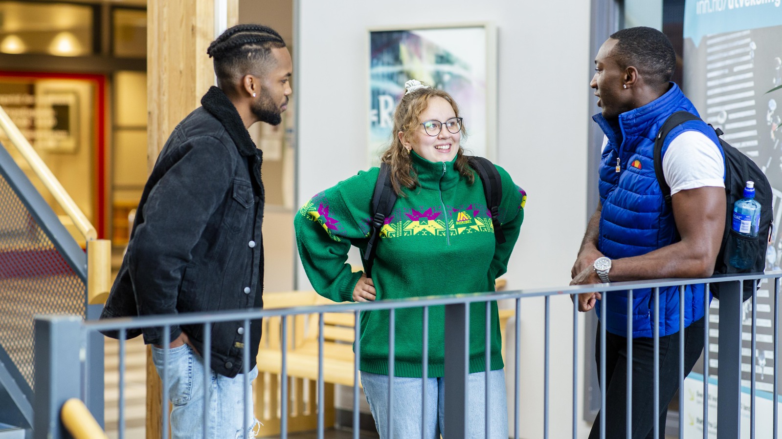 Tre studenter, to gutter og en jente står ved et gelender å snakker med hverandre. En har på seg mørk skjorte, en hvit t-shirt og blå vest, jenta har på seg grønn strikkegenser. 
