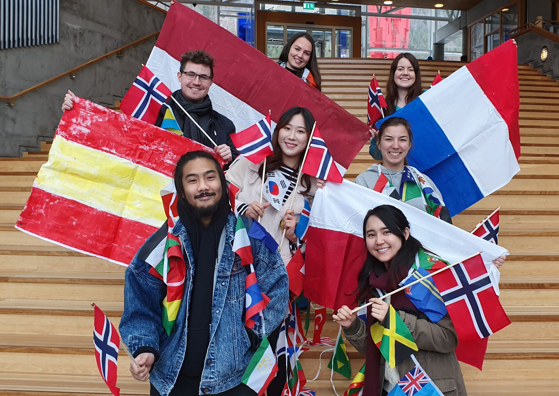 Internasjonale faddere står i en trapp, holder flagg fra ulike land og smiler