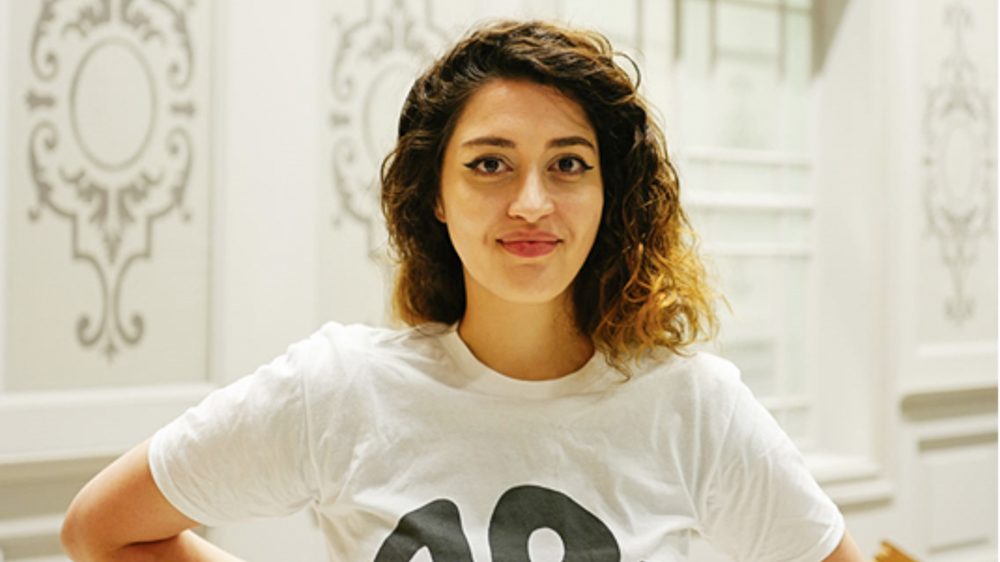 Portrettbilde av Nadja med hvit t-skjorte med svart skrift mot en hvit vegg med ornamenter