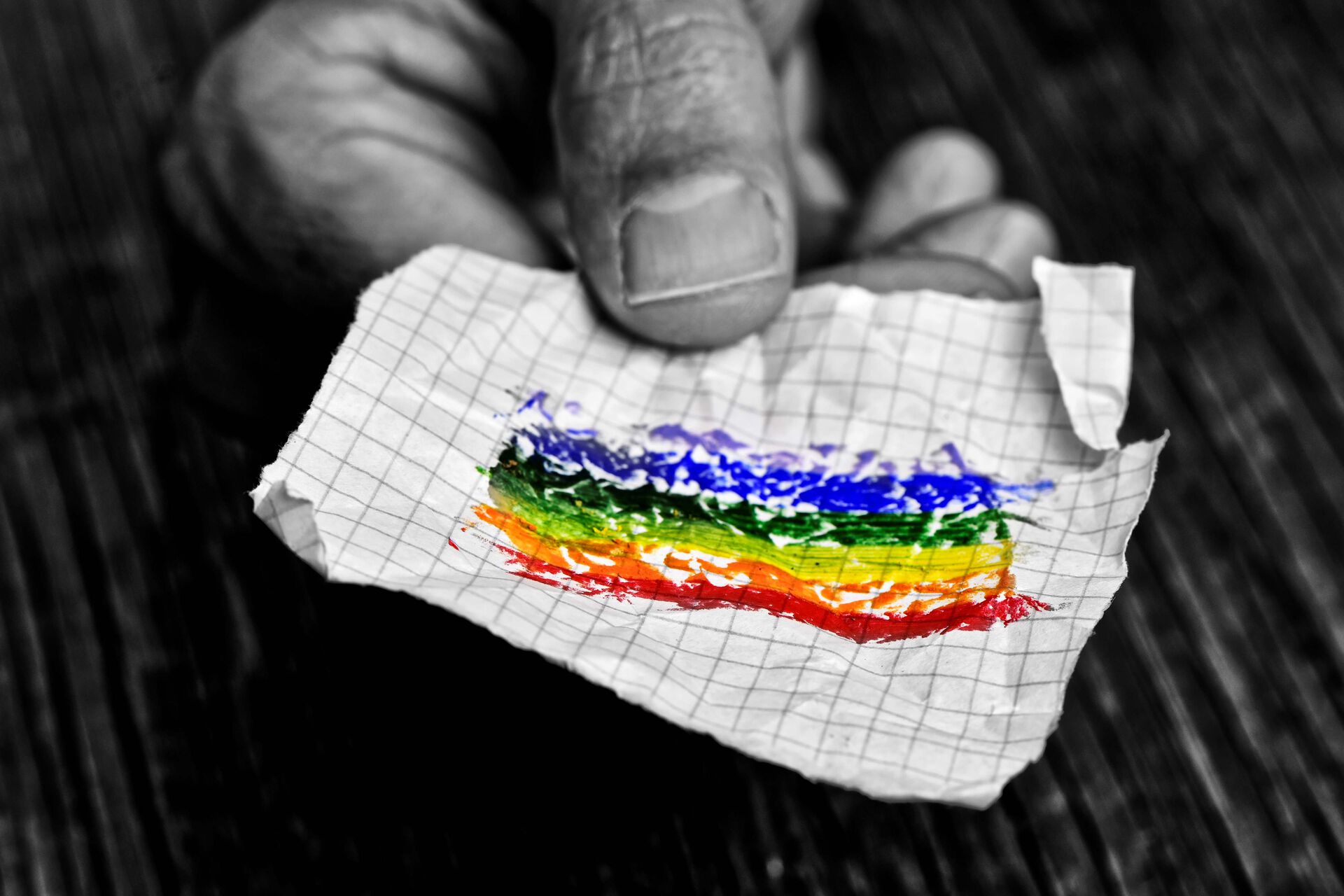 Sort-hvit foto av en hånd som holder fram en lapp med et håndtegnet regnbueflagg, lappen er i farger.