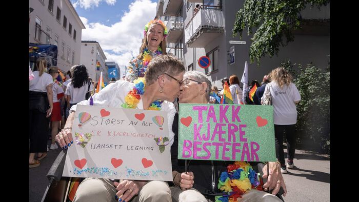 To eldre kvinner som sitter ved siden av hverandre og kysser hverandre. De holder plakater med tekst "Størst av alt er kjærligheten uansett legning" og "Takk bestefar". Prideparade og flere regnbueflagg i bakgrunnen.