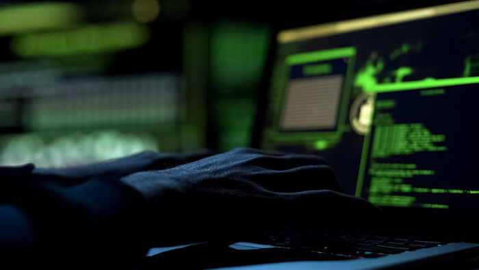 A dark room with a computer screen showing codes. We see the hands of a person working on the computer.