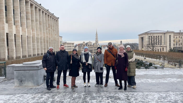 Eight people stand in a row, with their backs to the view of a view of a city. The weather is grey and there is a little snow on the ground.