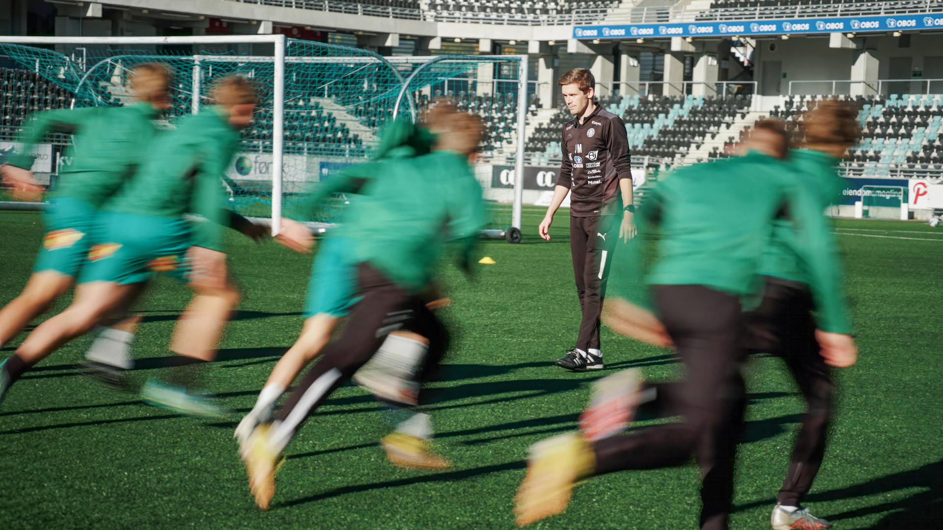 Mollatt i sort står midt inne i en flokk grønnkledde fotballspillere som løper over banen.