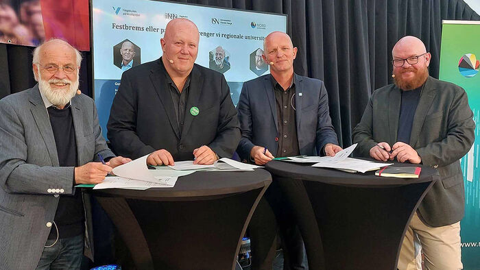 Fire menn i mørke skjorter som står bak et bord og signerer en samarbeidsavtale. 
