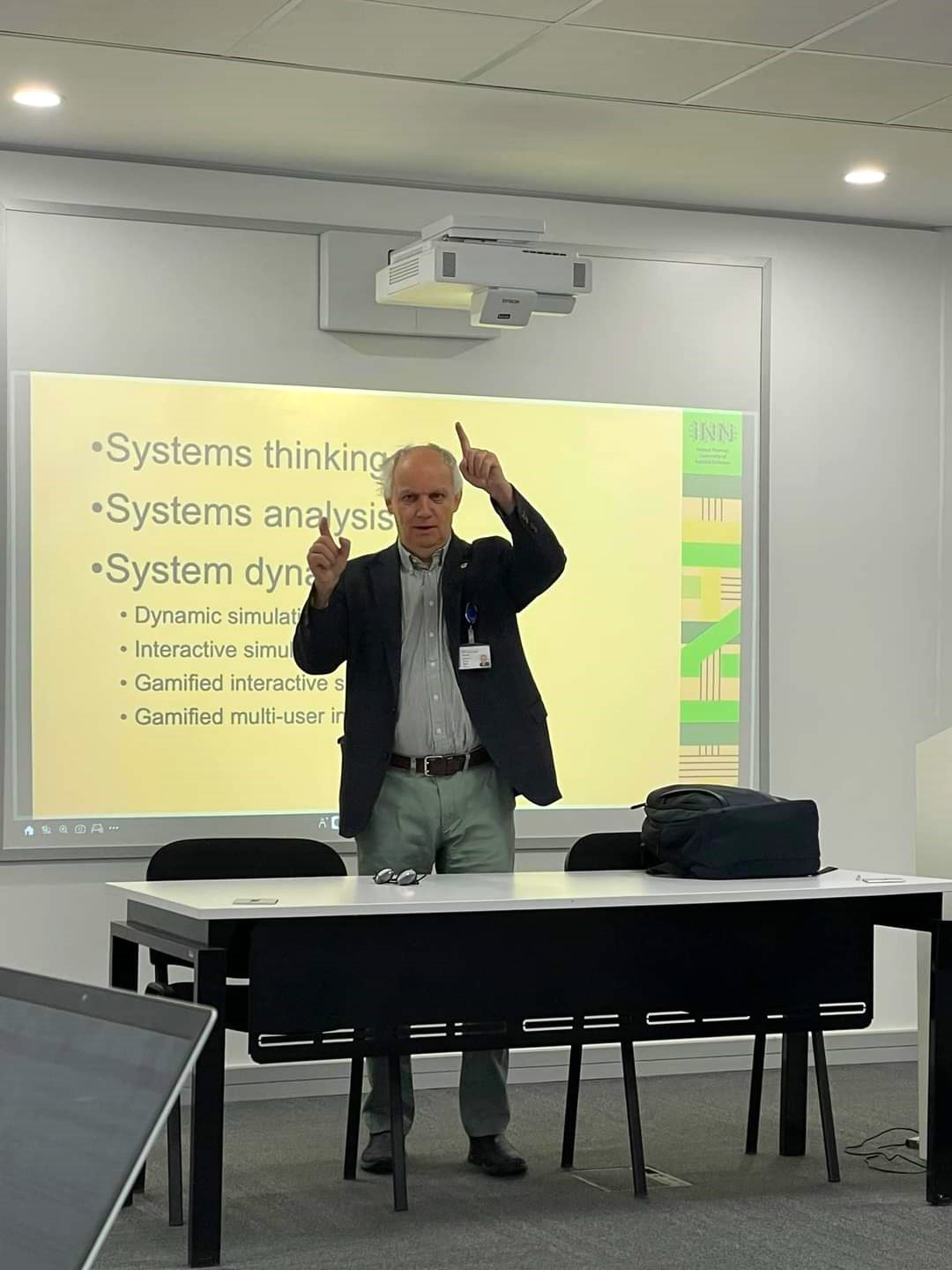 Professor Harald Sverdrup står bak et bord og gestikulerer, foran en storskjerm med med punktvis tekst om System thinking