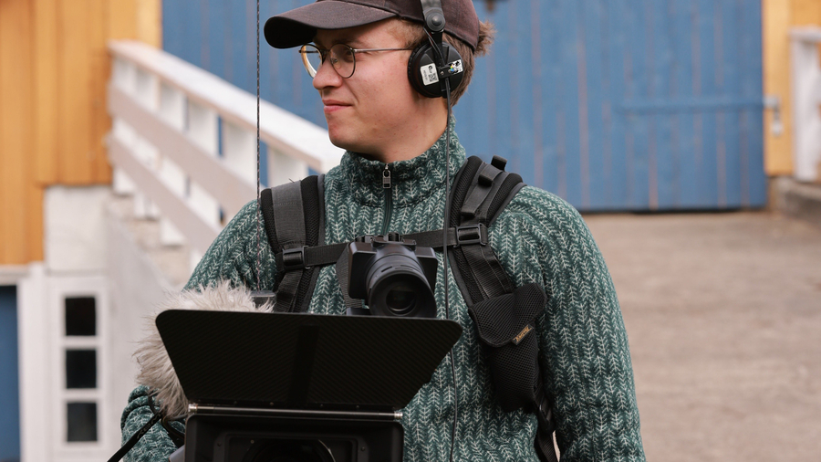 Åsmund Grini står med kamera- og lydutstyr foran en blå låvedør i grønn strikket genser. 