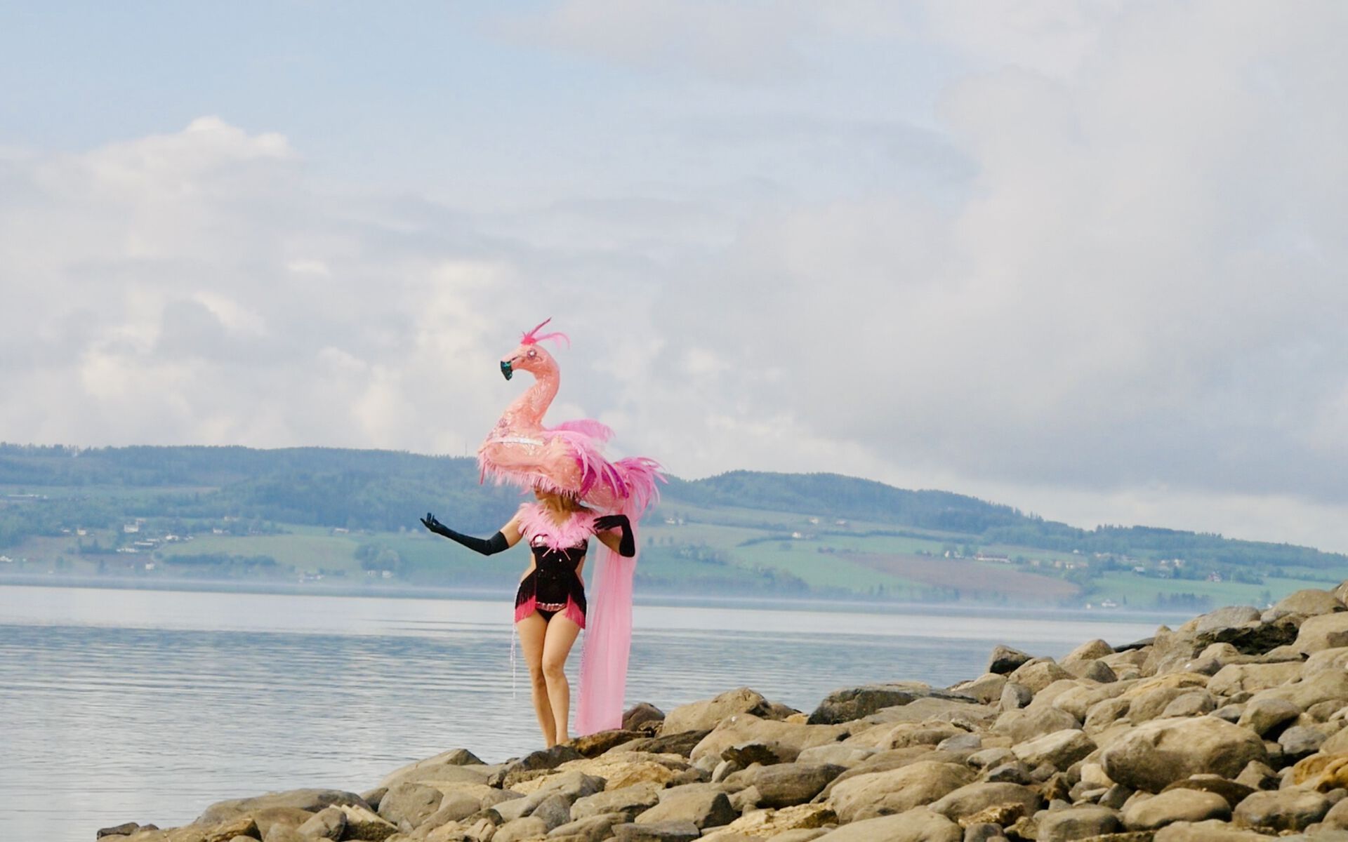 En dame i en svart og rosa badedrakt med  en rosa flamingo på hodet står på en en steinstrand foran et vann
