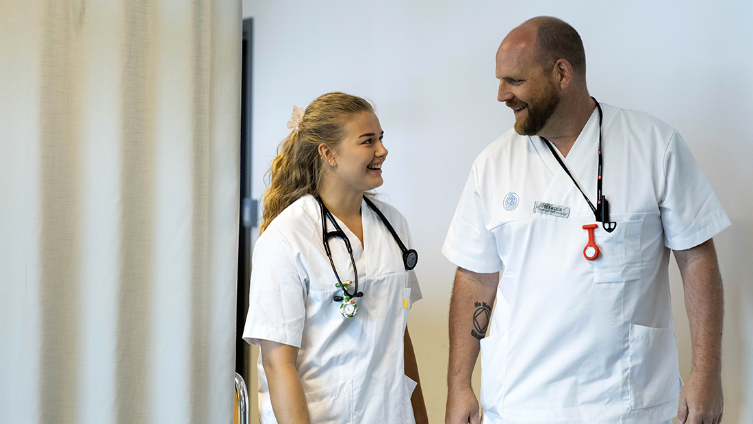 To sykepleiestudenter kledd i hvitt som smiler til hverandre. 