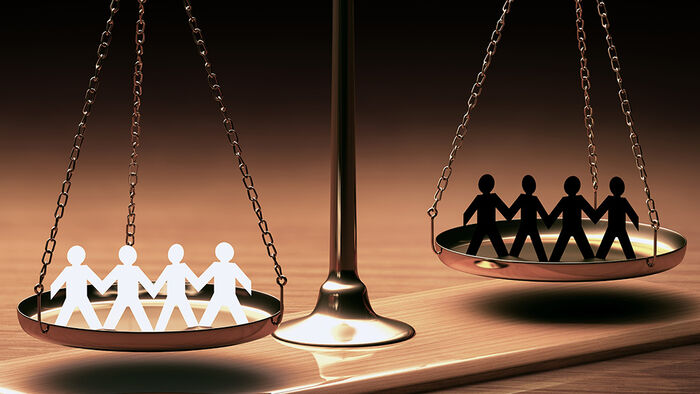Illustration photo showing balance scales with four white figures on the left side weighing more than four black figures on the right side.