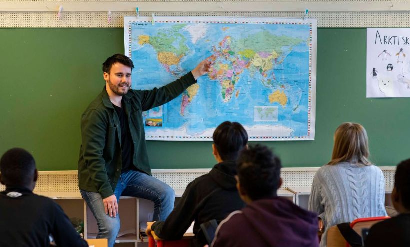 En lærer sitter foran en klasse og peker på et verdenskart.