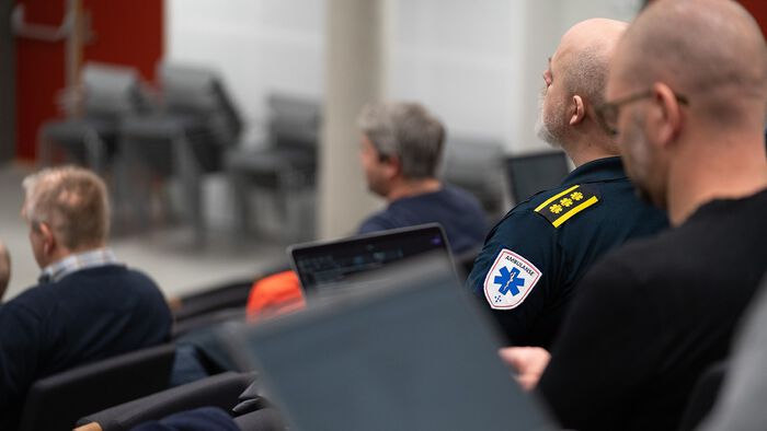 En gruppe mennesker sitter i et auditorium, en av dem har ambulanse-merke og tre stjerner på skulderen.