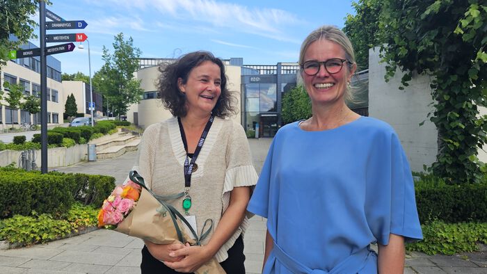 Bente Bolme and Stine Grønvold are standing in front of Hamar campus. They are smiling, and the sky is blue behind them.