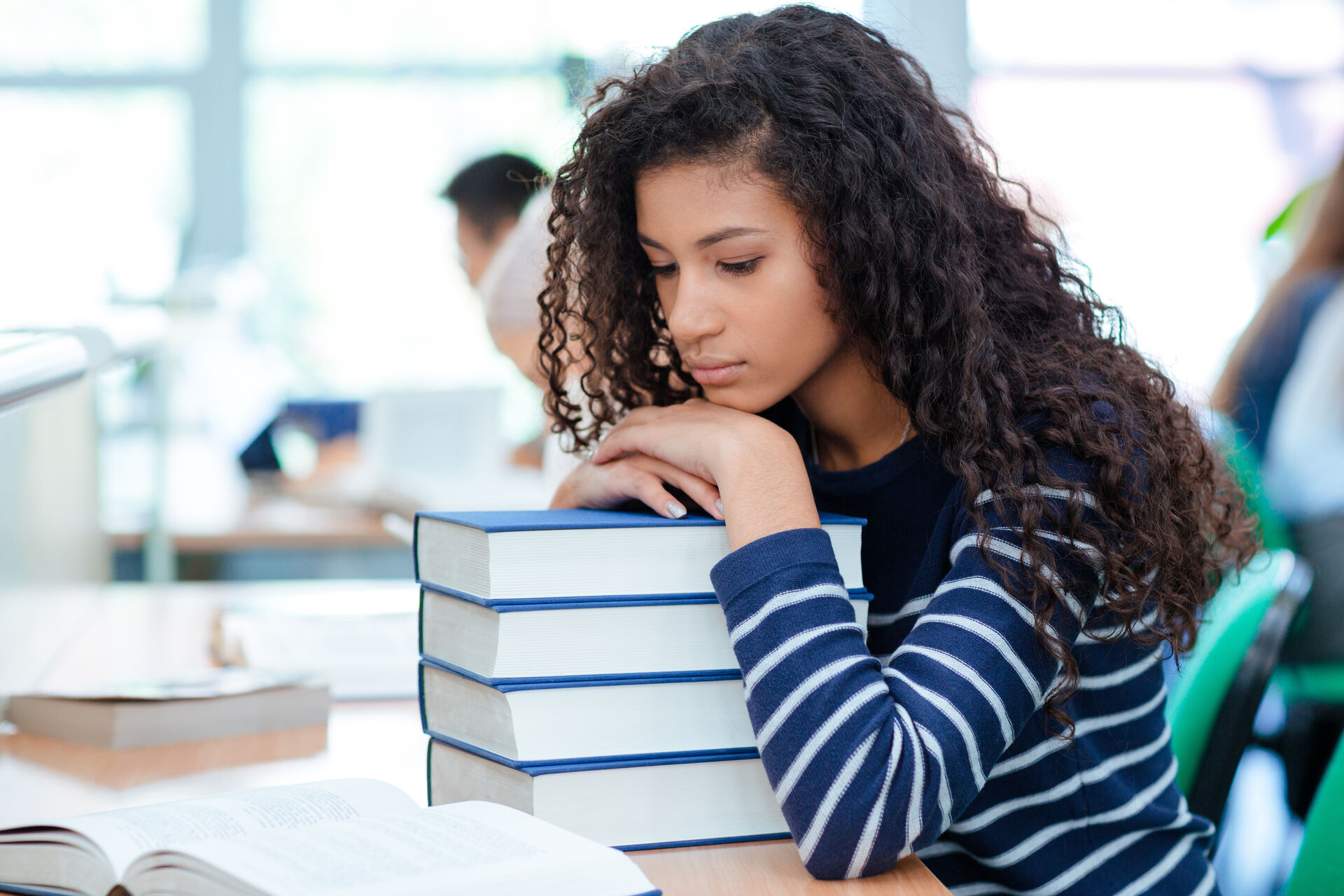 En kvinnelig student sitter på en lesesal og hviler hodet trist på en bunke med pensumbøker