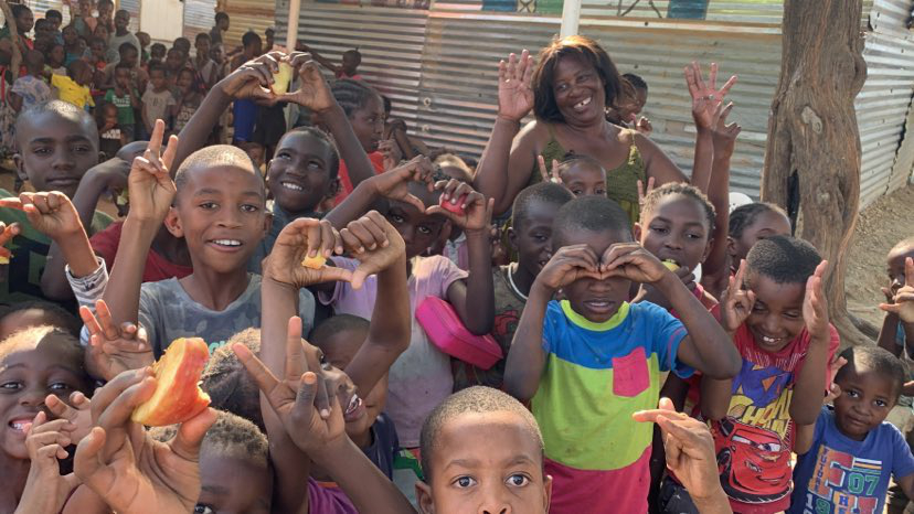 Mange glade skolebarn står samlet utenfor skolen sin i Nambia. De ser inn i kameraet mens de lager hjerter, fredstegn og andre hånduttrykk mot kameraet.