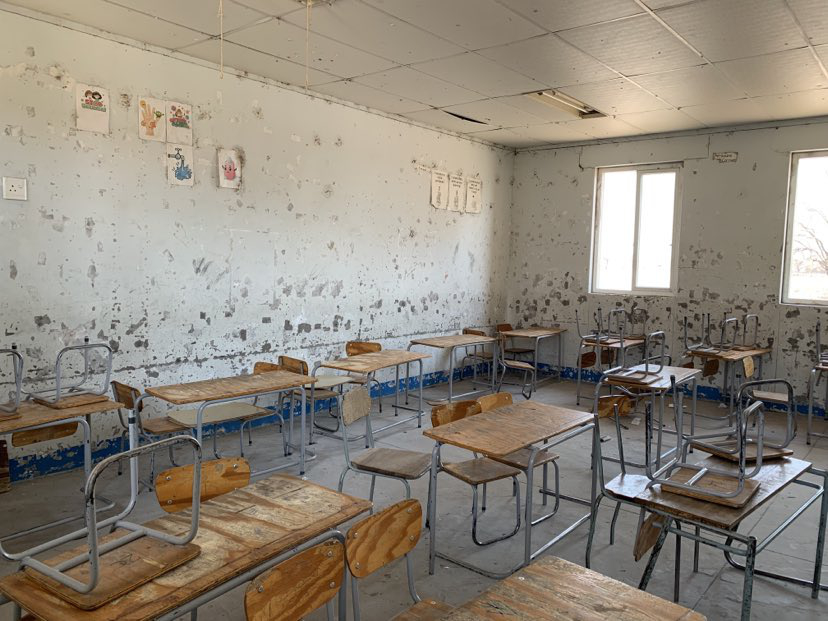 Et tomt, nedslitt klasserom med gamle pulter og stoler satt litt hulter til pulter. Det er en del tegningen hengt opp på den flekkede betongveggen i bakgrunnen.