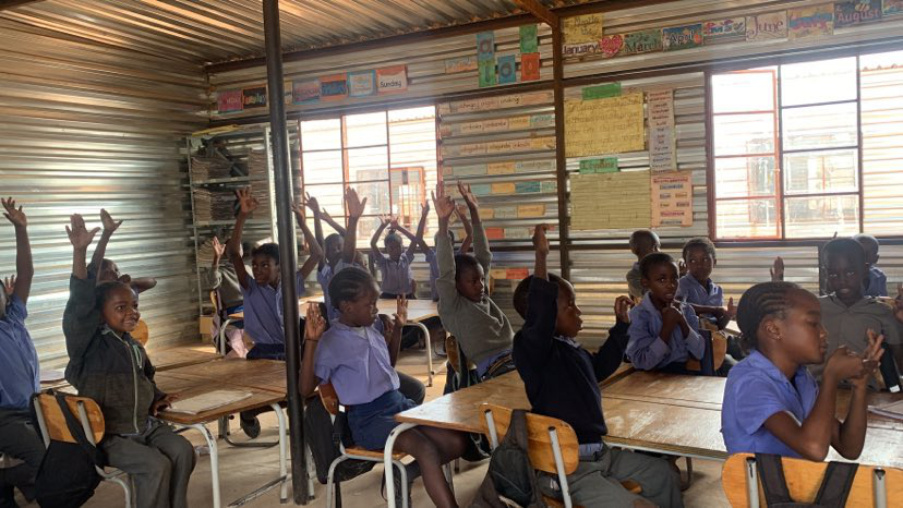 Mange namibianske skolebarn sitter i et klasserom og rekker opp hendene sine. Den ene blikkveggen bak er fylt med plakater og tegninger.