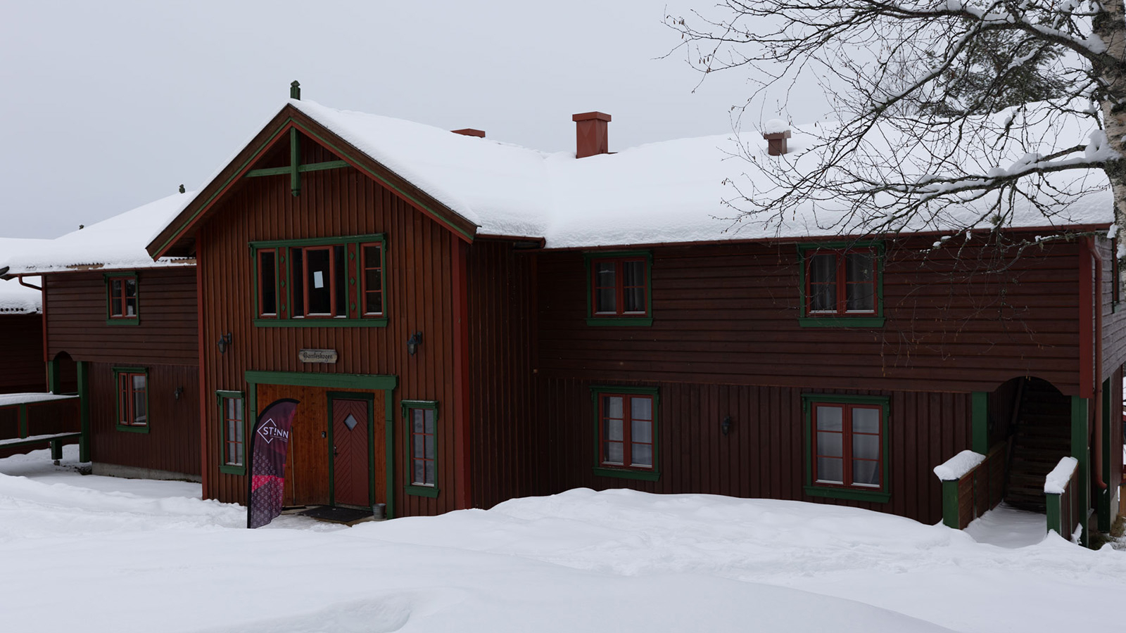 En stor hytte, dekket i snø, med et StInn-flagg utenfor hoveddøra.