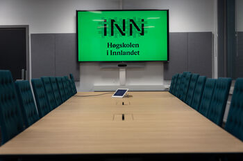 Et formelt møterom hvor identiske kontorstoler er satt pent inntil et stort konferansebord. I midten er en stor skjerm hvor Høgskolen i Innlandets logo blir vist.