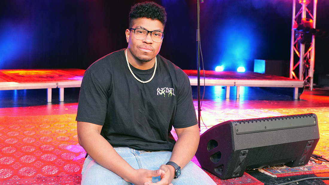 Mannlig musikkstudent i svart t-skjorte som sitter på en scene med sterke lys i bakgrunnen. 