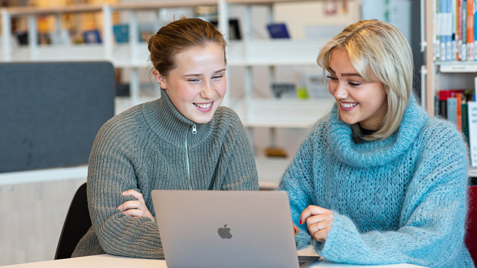 To kvinnelige studenter sitter fremfor en macbook og ser ned på skjermen. Den ene har på seg en grønn strikket ullgenser med glidelås, den andre en strikket turkis genser med høy hals.