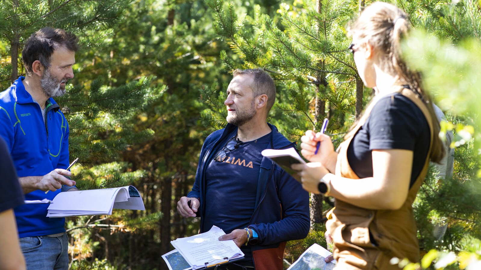 En mannlige ansatt snakker sammen med en mannlig og en kvinnelig student ute i skogen. Alle har papirer i hendene. De står i en furuskog.