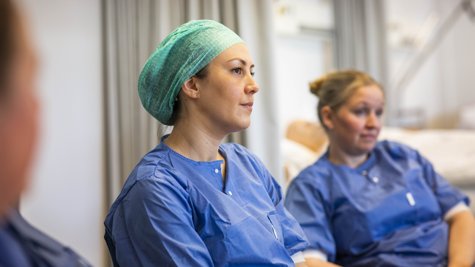 To operasjonssykepleiere ikledd blå operasjonsuniformer inkludert munnbind på seg i en operasonssal. Den ene justerer det blåteppet som henger over en arm som er synlig som skal opereres.