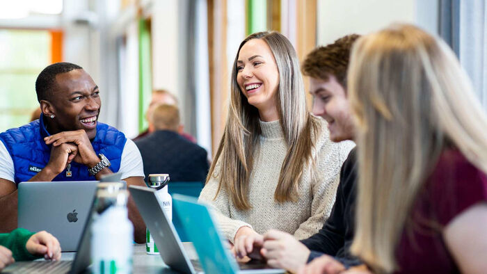 Fire smilende studenter med hver sin datamaskin snakker sammen ved et bord